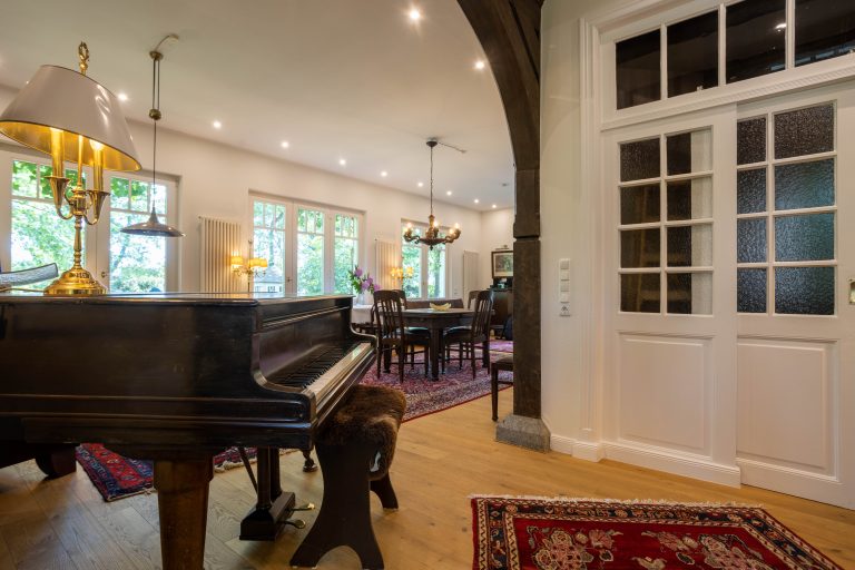 173A6513-HDR – Hubertus Boltenhagen Flügel im elegant beleuchteten Wohnzimmer mit Esstisch und großen Fenstern.