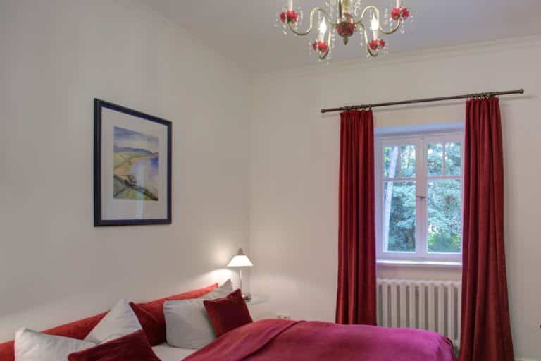 Haus_Hubertus_-12-2 (2) – Hubertus Boltenhagen Gemütliches Schlafzimmer mit roter Bettwäsche und Kristallleuchter.