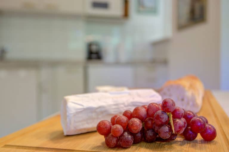 Haus_Hubertus_-62-5 – Hubertus Boltenhagen Rote Trauben und Brie auf einem Holzbrett in einer unscharfen Küche.