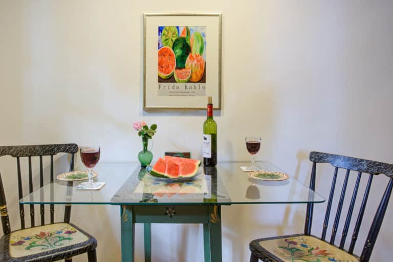 Haus_Hubertus_-7-11 – Hubertus Boltenhagen Glastisch mit Wein, Wassermelonen, Blumen und Stühlen; Wandbild im Hintergrund.