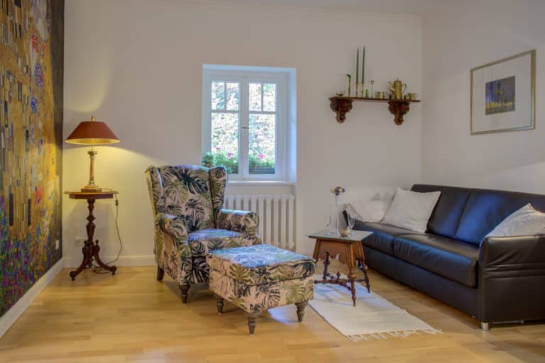 Haus_Hubertus_-7-20 – Hubertus Boltenhagen Gemütliches Wohnzimmer mit floralem Sessel, Sofa, Lampe und Fenster.