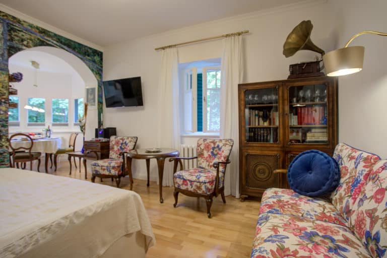 Haus_Hubertus_-7-23 – Hubertus Boltenhagen Helles Wohnzimmer mit antikem Grammophon, Holzschrank und blumigen Sesseln.