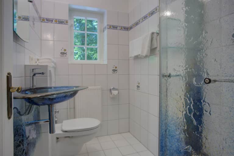 Haus_Hubertus_-72-4 – Hubertus Boltenhagen Helles Badezimmer mit Fenster, weiß gefliest, Glasdusche und Wand-WC.