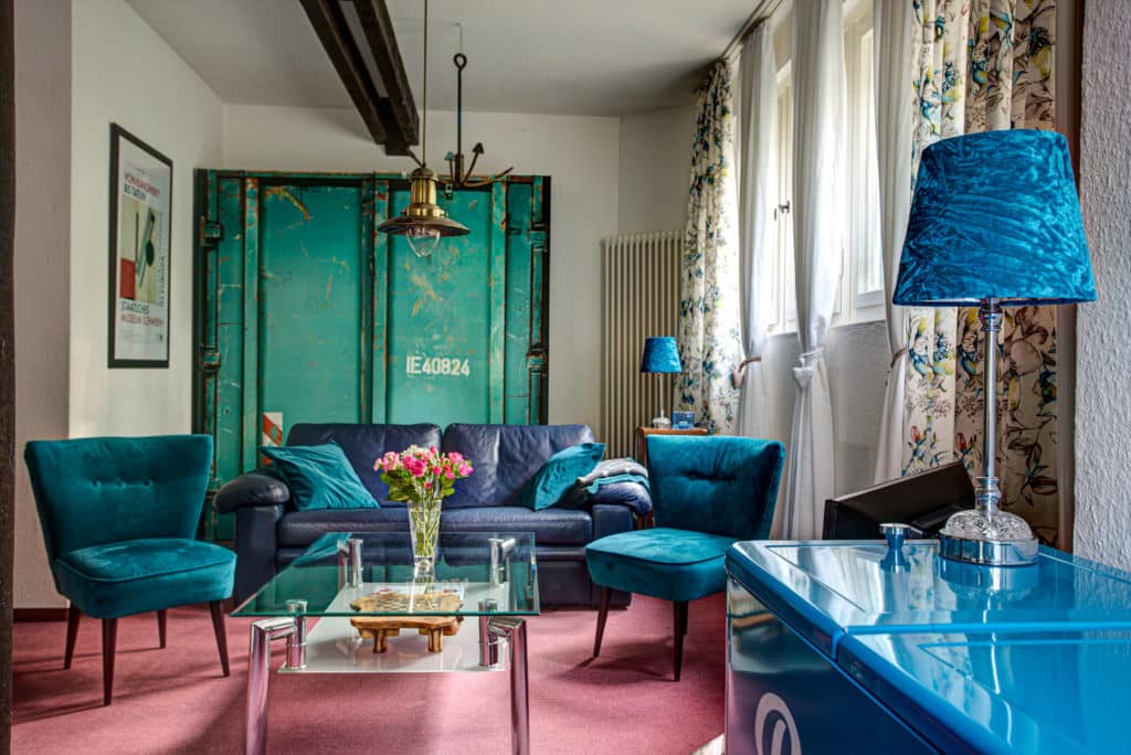 Hubertus7-93 – Hubertus Boltenhagen Wohnzimmer mit blauen Möbeln, rosa Teppich, Blumen auf Glastisch.
