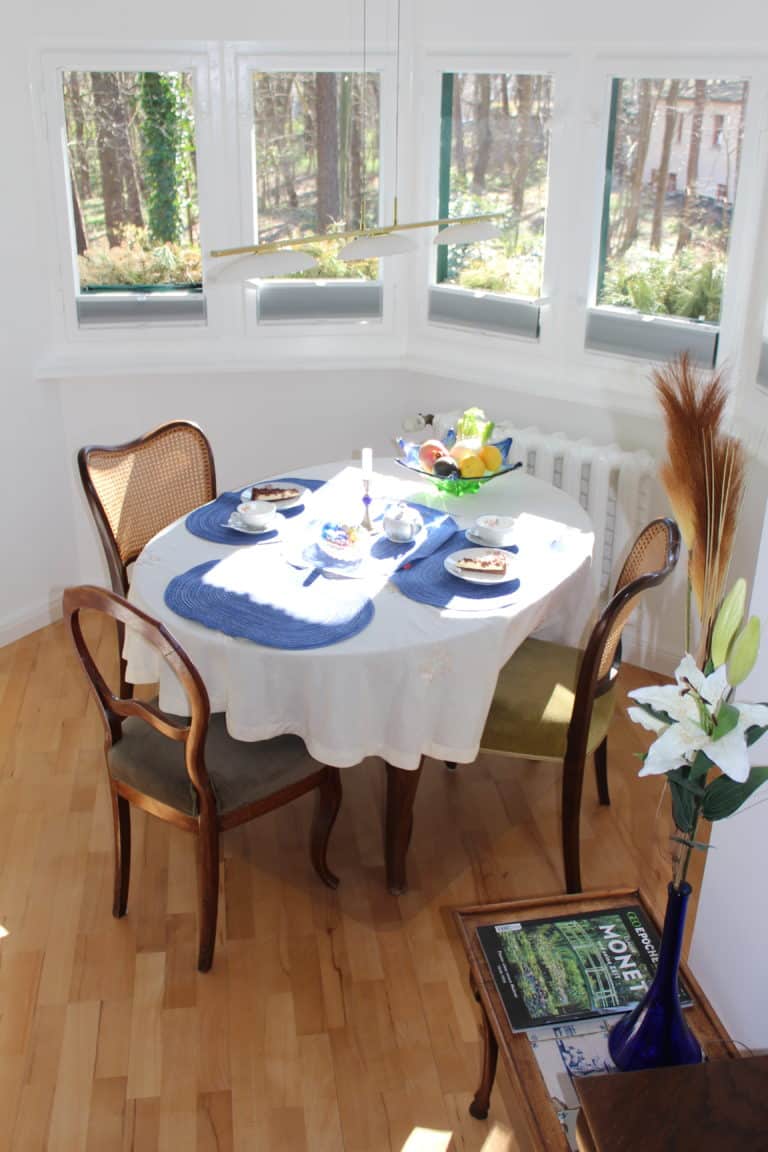 IMG_5909 – Hubertus Boltenhagen Runder Tisch mit weißen Decken, blauen Platzsets und Holzmöbeln in einem sonnigen Raum.