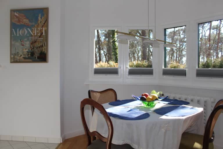 IMG_5923 – Hubertus Boltenhagen Runder Tisch mit blauer Tischdecke, Früchteschale, Blick auf Wald durch Fenster.