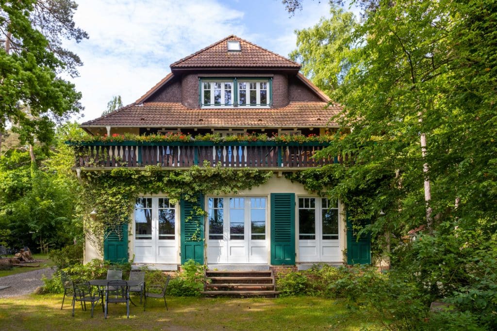 HausHubertus_Aussen-0889 – Hubertus Boltenhagen Zweistöckiges Haus mit grünen Fensterläden, umgeben von Pflanzen.