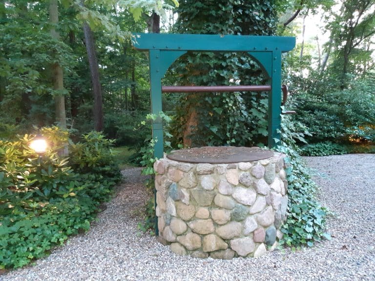 20200717_212849 – Hubertus Boltenhagen Stone fountain in the forest, surrounded by a gravel path and green vegetation.