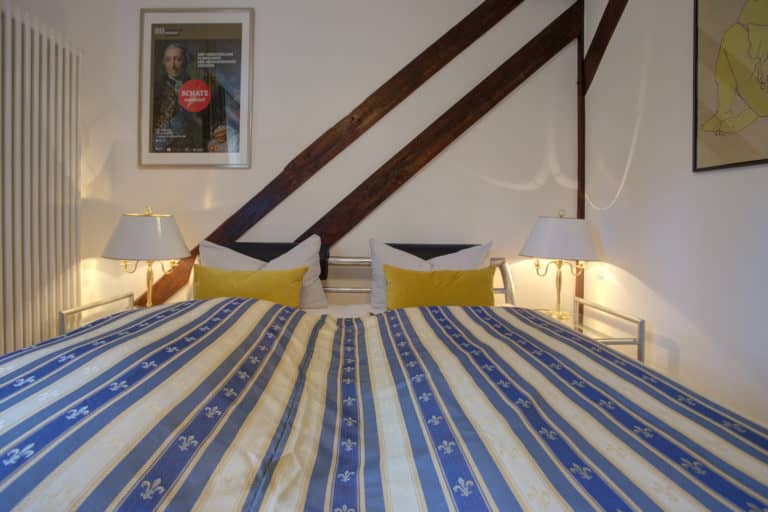 Haus_Hubertus_-92-4 – Hubertus Boltenhagen Bed with striped blanket, yellow pillows and slanted wooden beams on the wall.