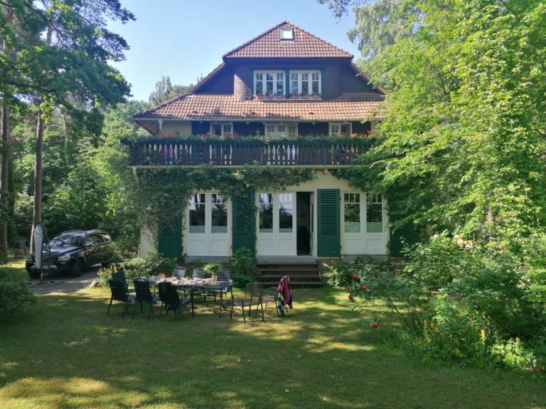 IMG-20210707-WA0000 – Hubertus Boltenhagen Dreistöckiges Haus mit Balkon, umgeben von Bäumen und Gartenmöbeln im Vordergrund.