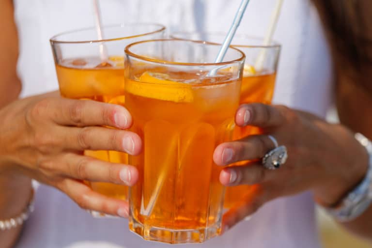 _79A8858 – Hubertus Boltenhagen Three glasses of orange juice with ice cubes, held in hands.