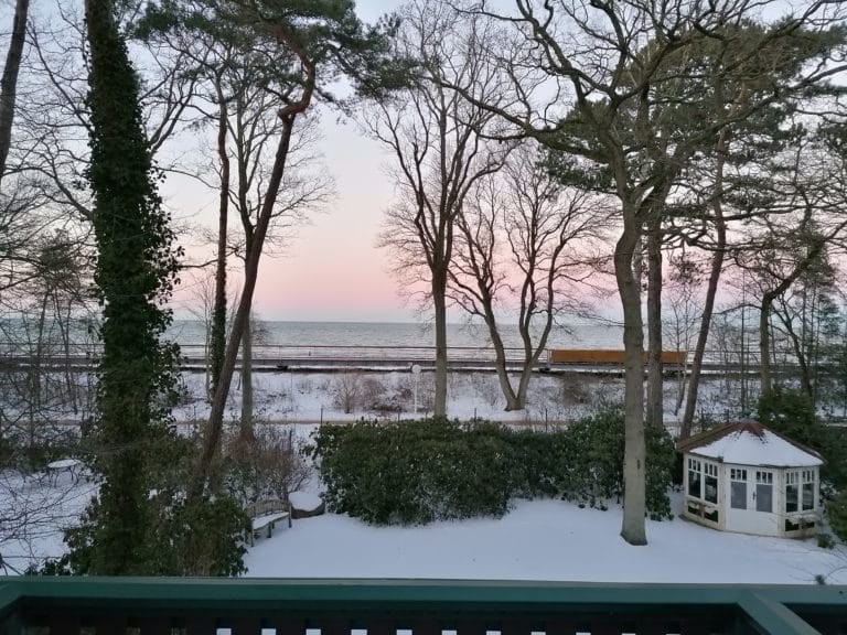 IMG_20210212_172307 – Hubertus Boltenhagen Snow-covered garden with trees against a pink sky and sea in the background.