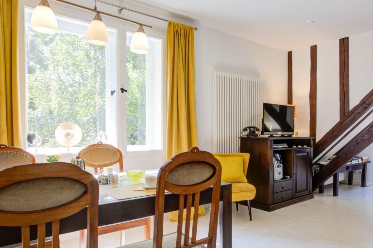 Haus_Hubertus_-402 2 – Hubertus Boltenhagen Esszimmer mit Holztisch, gelben Vorhängen und Sessel, TV-Schrank.
