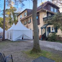 IMG-20260428-WA0006 – Hubertus Boltenhagen Hinteransicht einer Strandvilla mit zwei weißen Zelten im Garten, umgeben von Bäumen und einem Kiesweg.