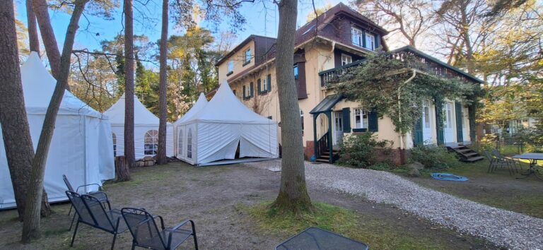 IMG-20260428-WA0006 – Hubertus Boltenhagen Rear view of a beach villa with two white tents in the garden, surrounded by trees and a gravel path.