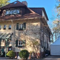 IMG-20260428-WA0010 – Hubertus Boltenhagen Strandvilla mit brauner Fassade, grünen Fensterrahmen und einem Zelt auf dem Grundstück, umgeben von Bäumen.